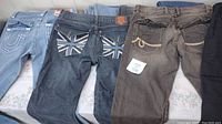 Photo showing three pairs of jeans: light blue True Religion style, dark denim with Union Jack design, brown jeans with rope pocket accents.