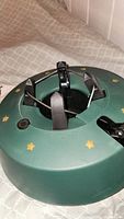 Close-up photo of green Christmas tree stand featuring black locking foot lever and gold star decorations on top surface.