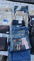 Photo showing a selection of women's and men's jeans and slacks spread out on a floral patterned surface, featuring denim jeans with embroidery and various beige and blue pants.