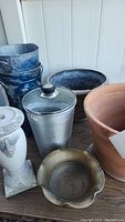 Lot items on a wooden surface showing scalloped edge gold bowl, large terracotta planter, three metal pails (one with lid), blue speckled metal buckets, grey urn-style planter, and a black container.