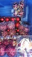 Various bags of red and gold blown glass Christmas ball ornaments, some with glitter and striped patterns, in stacked plastic packaging inside a storage tub.