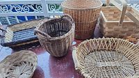 Photo showing six unique woven baskets of various sizes and shapes on a red table. Includes baskets with integrated handles, a square basket with swing handles, a tall cylindrical waste basket, and others.