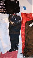 Top-down photo of seven pairs of pants laid flat, showing various colors and embroidered patterns including black, white, red, and green colors.