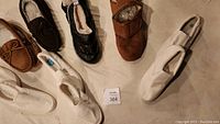 Seven pairs of shoes displayed on floor including brown suede moccasins, dark brown moccasins with fur lining, black poofy slip-ons, brown Velcro closure slip-ons, and multiple white canvas slip-ons.