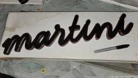 Wood carved wall sign with the word 'martini' shown on a white surface, with a marker nearby for scale.