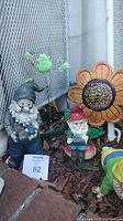 Photo showing a large garden gnome with blue hat and pipe, a smaller gnome sitting on mushroom, a green frog, and parts of metal flower yard spikes in the background.