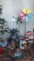 Various garden statuaries and yard art pieces arranged in mulch including frogs, fish, bears, gnome, and flower pinwheel.