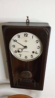 Full view of Alkosha wall clock mounted on wall showing front wooden case, clock face, pendulum and glass door.