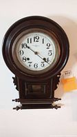 Full front view of antique wall clock showcasing dark wood frame, white clock face with black numerals, and protective glass door.