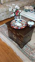 Small wooden Asian inspired table with storage drawers, brass hardware and handles, placed on a patterned rug with floral upholstery sofa in background. On top are three Asian inspired ceramic dishes and one teacup, along with a floral arrangement in a ceramic vase.