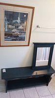 Framed artwork print above dark wooden bench with matching framed mirror leaning on bench.