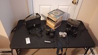 Wide shot displaying the Canon and Minolta cameras, Scotch film cartridge, binoculars, stacked film boxes, and part of the movie camera and case on a black table.