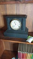 Wooden black mantel clock with Roman numerals, closed glass cover, placed on wooden shelf.