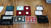Overview photo of all coin sets and commemorative coins arranged on wood surface showing the different packaging and coins included.