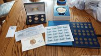 Photo showing multiple coin collections displayed on a wooden table including a blue Lincoln Head Cent collection holder, 24Kt gold plated set in presentation box, stamp sheet, and some paperwork.