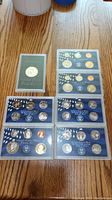 Photo showing seven United States Mint proof coin sets in plastic cases arranged on a wooden surface.