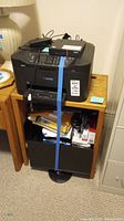 Canon MAXIFY printer with blue tape securing it on a wooden cabinet containing papers and electrical cords.