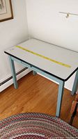 White enamel topped table with black edging, painted blue wooden base and legs on hardwood floor next to framed artwork.