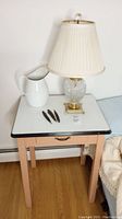 Full view of enamel topped table with drawer, glass lamp, enamelware pitcher, and three vintage straight razors.