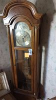 Full view of the grandfather clock in wooden case, glass front door showing pendulum and weights beneath clock face.