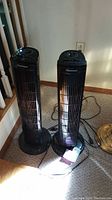 Two Kenmore tower fans standing side by side on carpet near wall outlet.