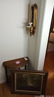 View of oil painting leaning against wall with half-moon wood table and gold wall hanging sconce above
