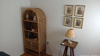 Wicker shelf with items displayed on shelves, wooden side table with lamp and framed artwork on wall behind