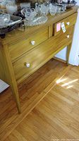 Wooden sideboard with three drawers and white knobs shown standing on hardwood floor with glassware on top.
