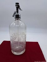 Full view of the glass seltzer bottle showing etched logo and metal siphon top on red background.