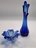 Two blue art glass pieces shown side by side on a white background: a ruffled edge dish and a tall ruffled edge vase.