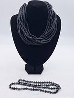 Full view of multi-strand bead necklace on black necklace form with separate single-strand spherical bead necklace lying in front.