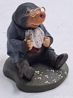 Full view of the handpainted mole figurine seated on a textured base, wearing glasses, bib, and jacket.