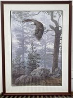Full view of the framed bald eagle print showing the eagle flying over pine trees and rocks in a muted color palette with a brown wooden frame.