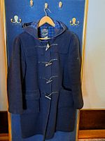 Full front view of navy blue Gloverall duffel coat hung on hanger against a blue backdrop with gold hooks.