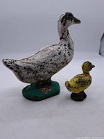 Two painted concrete ducks, one larger white with green base and one smaller yellow with red and blue accents, showing wear and paint chipping.