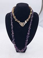 Two gemstone necklaces on black bust display: one short multi-colored polished stone necklace with a central fan-shaped pendant and one long purple amethyst chip bead necklace.