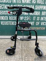 Full view of the black Nexus folding walker showing its three wheels, padded seat, handlebars with handbrakes, and folding frame.