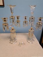Full view of two tall antique glass candelabra with candle holders and hanging crystal prism drops, placed on a table.