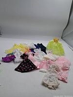 Top-down view of various vintage Barbie doll outfits laid on a flat surface showing diverse colors and styles including dresses and skirts.