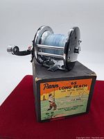 Fishing reel on top of original box. Shows front and handle side of reel, spool filled with blue fishing line.
