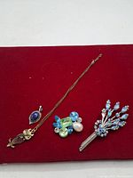 Photo of a vintage gold-tone necklace with blue oval pendant, a cluster brooch with blue and green stones plus a faux pearl, and a floral brooch with blue rhinestones on a red background.