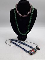 Three vintage necklaces displayed on black jewelry bust: green stone bead necklace, purple shell bead necklace, and blue beaded necklace with colorful pendant