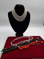 Multi-strand faux pearl necklace on black display bust with three other beaded necklaces laid on red velvet cloth.