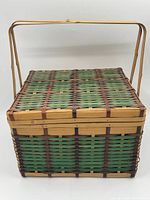 Front view of a vintage wicker picnic basket showing green, natural, and brown woven wicker with wooden handle and flat hinged lid.