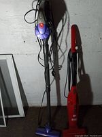 Two stick vacuums standing upright next to each other on floor, one purple/black Dirt Devil with red button, one red Gravitti, cords visible.