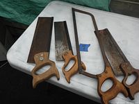 Photo shows three back saws and one hack saw arranged on white surface; saws have wooden handles and metal blades with visible rust and wear.