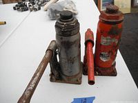 Two older hydraulic bottle jacks, one silver and one red, both showing significant wear and rust. Shown with pumping handle detached and resting alongside.