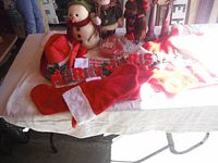 Photo showing several Santa hats, two Christmas stockings—one red and one plaid, 'MERRY CHRISTMAS' sign, stuffed moose, musical snowman, and other Christmas figurines on a white fabric surface.