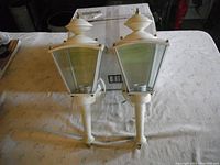Pair of white coach lights with pointed finial tops and clear glass panels. Mounted on tapered arms, placed on a white blanket with a cardboard box in the background.