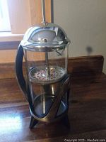 French press coffee/tea maker side angle showing clear double wall glass and metal frame base with black handle.